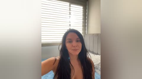 Media: A video of a young woman with long, dark hair, fair skin, and a slim physique, standing in a bright bedroom with white blinds partially open.