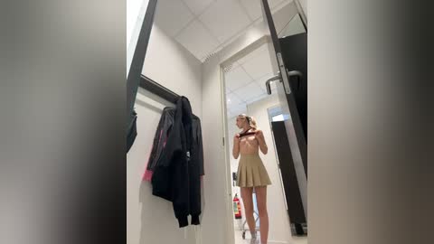 Media: Video of a woman in a dressing room, wearing a beige skirt and holding a black jacket. She stands in front of a full-length mirror, reflected in the mirror's glass. The room has white walls and a tiled floor.