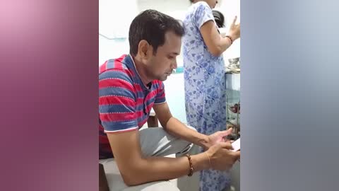 Media: Video of a South Asian man in a red and blue striped t-shirt, sitting on a bed, adjusting his smartphone. A woman in a blue floral-patterned hospital gown is standing behind him.