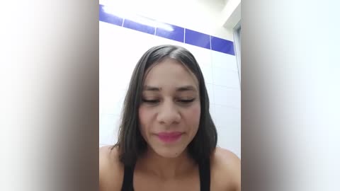 Media: Video of a young Latina woman with straight, shoulder-length brown hair, wearing a black tank top, looking down with a neutral expression in a tiled bathroom with blue and white tiles.