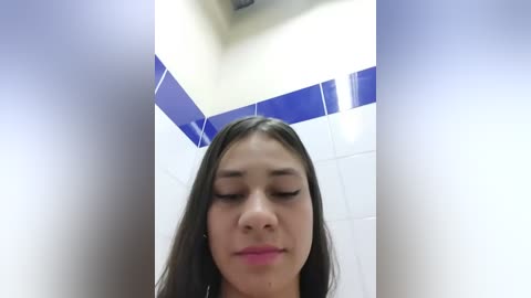 Media: Video of a young Latina woman with long, straight brown hair, fair skin, and closed eyes, standing in a tiled bathroom with white and blue accents.