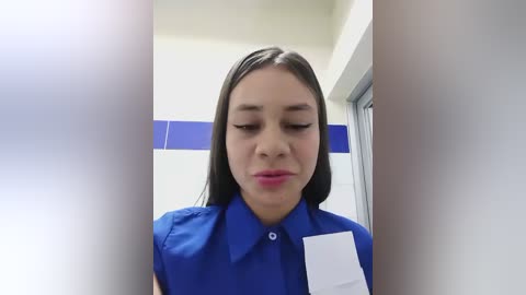 Media: Video of a young woman with straight black hair, wearing a blue uniform shirt with a white name tag, standing in a brightly lit bathroom with white and blue tiled walls.