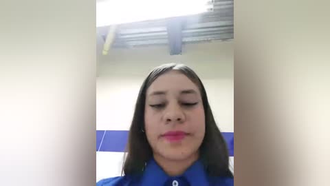 Media: Video of a young Latina woman with medium brown skin, long dark hair, and pink lipstick, wearing a blue shirt, taken indoors with a white ceiling and blue tiled wall background.