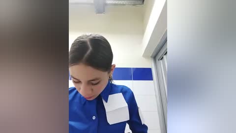 Media: Video of a young woman with dark hair in a ponytail, wearing a bright blue uniform with a white pocket, standing in a white-tiled corridor with blue accents and a frosted glass door.