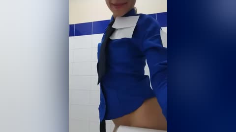 Media: Video of a woman in a blue blazer with a white collar and black tie, partially unzipped, revealing a white bra and no pants in a white-tiled bathroom with blue trim.