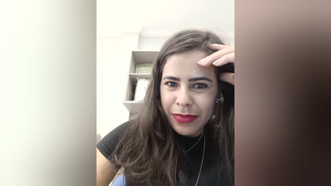 Media: Video of a Latina woman with long dark hair, wearing a black top and red lipstick, taking a selfie in a room with a beige wall and bookshelf.