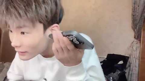 Media: Video of a young Asian boy with short brown hair, wearing a white shirt, trimming his ear hair with a small black razor. Background shows a plain beige wall and a cluttered room with a wooden door.