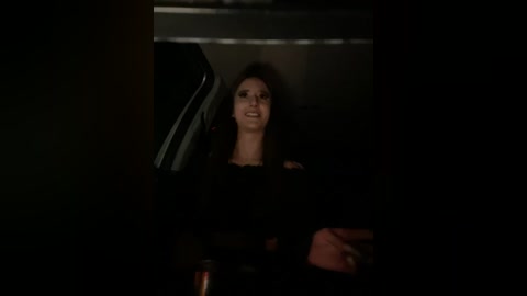 Media: A dimly-lit video of a woman with dark hair and fair skin, smiling, wearing a black dress, sitting in a dimly-lit car interior.