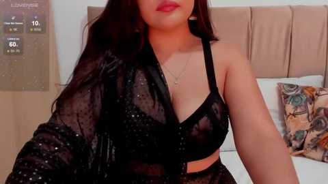 Media: Video of a woman with long black hair, wearing a sheer black polka-dot blouse revealing a black bra, sitting on a bed with beige headboard.