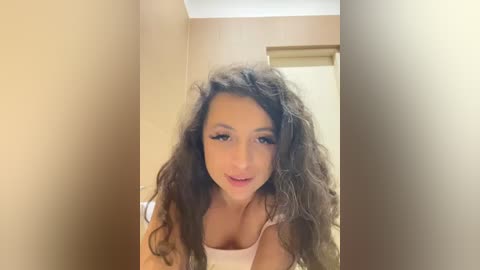 Media: Video of a young woman with curly, dark brown hair and light skin, wearing a white tank top, standing in a beige hallway with a wooden door in the background.