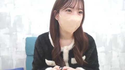 Media: A video of an Asian woman with long brown hair, wearing a black fur-trimmed coat, white mask, and gold hoop earrings, sitting in a brightly lit room with a blurred background.