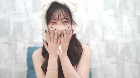 Media: Video of a young Asian woman with long dark hair, wearing a fluffy white hat, holding her hands over her face, against a soft, light-colored, textured background.