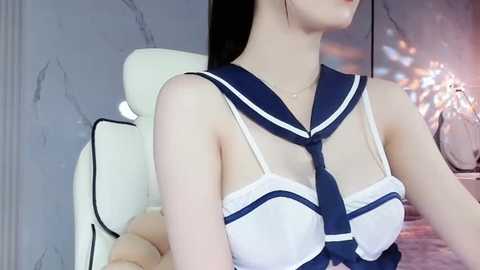 Media: Video of a slender, fair-skinned woman with long black hair, wearing a sheer, white sailor-style top with a navy blue collar and bow. She sits on a white gaming chair, in a dimly lit, modern room with gray marble walls.