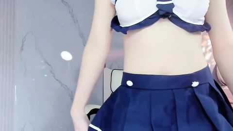 Media: Video of a slim, fair-skinned woman wearing a white and navy blue sailor-style bikini top and matching pleated skirt. The background features a light gray marble wall.