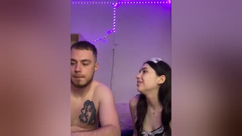 Media: Video of a shirtless man with a tattoo on his chest, standing beside a woman with long brown hair and a white headband, in a dimly lit room with purple LED strip lights.