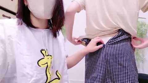 Media: Video of two women adjusting their clothing: one in a white shirt with a cartoon kangaroo graphic, the other in a beige shirt and black checkered skirt.