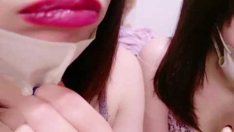 Media: Close-up video of a woman with long, straight black hair, wearing a light-colored top. Her lips are vivid pink, and she appears to be leaning forward, with her hand on her thigh. The background is blurred, focusing on her face and upper body.