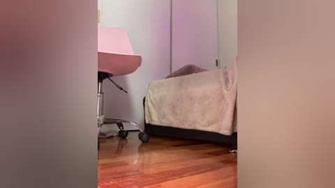 Media: A video of a minimalist bedroom with a single bed covered in beige bedding and a pink office chair with a white lampshade. The wooden floor and pale walls contribute to a serene, uncluttered ambiance.