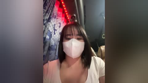 Media: Video of an Asian woman with short black hair, wearing a white face mask, sitting in a dimly lit room with blue and red LED lights.