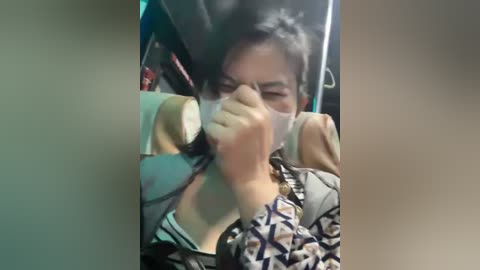 Media: A video shows an Asian woman with dark hair, wearing a patterned jacket, coughing into a tissue on a bus. The background is blurry, with a green and beige interior.