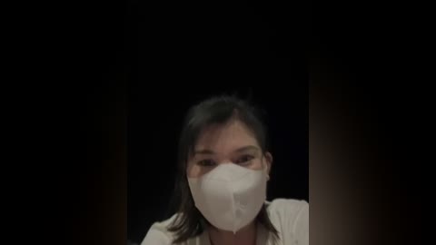 Media: Video of an Asian woman with dark hair, wearing a white face mask, against a black background, looking directly at the camera.