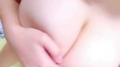 Media: Video of a fair-skinned person's bare breast, with their hand cupping and gently pressing the breast, creating a soft, natural texture. The background is blurred, focusing on the intimate, close-up detail.