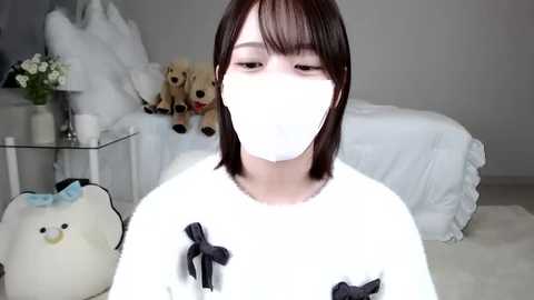 Media: A video of a young Asian woman with shoulder-length brown hair, wearing a white mask, white robe with black bows, and seated in a minimalist bedroom with plush toys, a bed, and flowers.