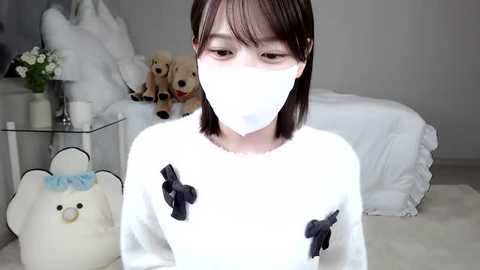 Media: A video of a young East Asian person with short brown hair, wearing a white face mask, a white sweater with black bows, in a minimalistic, white-themed bedroom with teddy bears and a glass table.