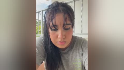 Media: Video of a young woman with wet, dark hair, wearing a grey T-shirt, looking down, on a balcony with a white railing and green foliage in the background.