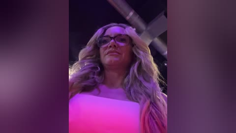 Media: Video of a blonde woman with long, wavy hair, wearing black glasses and a white top, standing in a dimly lit room with purple and pink lighting.