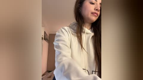 Media: Video of a young woman with long brown hair, wearing a white hoodie, standing indoors, smiling slightly, looking right. Background features neutral-toned walls and a door.