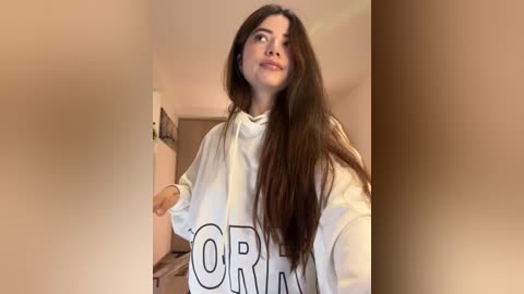 Media: Video of a young woman with long brown hair, wearing a white hoodie with \"ORANGE\" in black letters. She is indoors, standing near a beige wall, with a neutral expression.