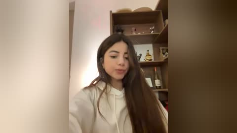 Media: Video of a young woman with long dark hair, wearing a white blouse, standing indoors. Background shows a wooden bookshelf with books, statues, and decorative items.