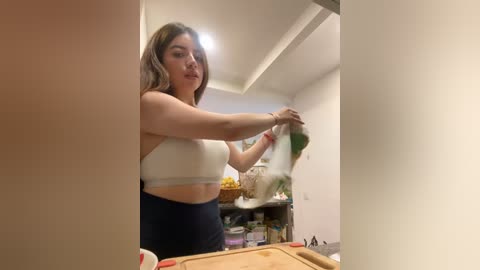 Media: Video of a young woman with long, wavy hair in a white sports bra and high-waisted black leggings, cleaning a glass door with a green cloth in a modern kitchen.