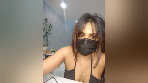 Media: Video of a young woman with medium skin tone, wearing a black face mask and black tank top, sitting in a dimly lit room with a plant and cluttered background.