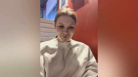 Media: Video of a woman with light skin and short brown hair, wearing a white surgical gown, standing in a brightly lit, modern hallway with red and orange walls and white horizontal blinds.