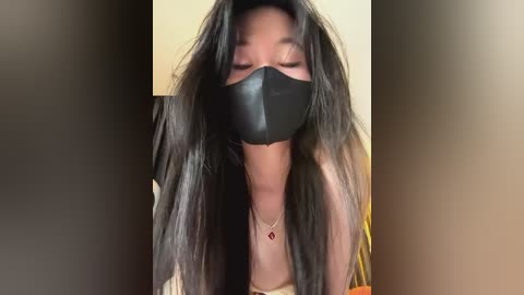 Media: Video of a woman with long, straight black hair, wearing a black face mask and a beige top, looking downward. Background is blurred, featuring beige and brown tones.