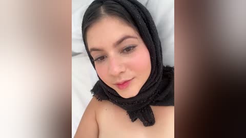 Media: Video of a young woman with fair skin and dark hair, wearing a black, fringed headscarf, lying on a white bed, smiling softly.