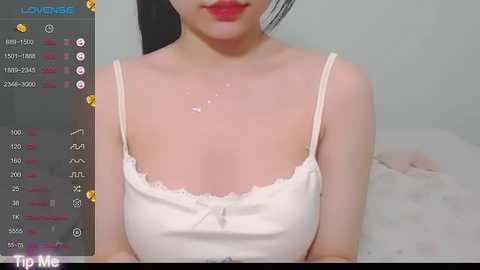 Media: Video of an East Asian woman with fair skin, wearing a white camisole, sitting on a bed with a white sheet. Her face is partially visible with red lipstick.