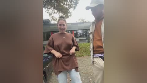 Media: Video of a woman in a brown, oversized shirt, with a smiling face, standing outdoors near a building with a green roof.