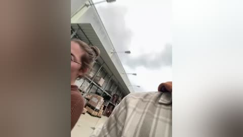 Media: Video of a woman with glasses, wearing a brown sweater, inside a warehouse with metal shelves, cloudy sky overhead, and a man in a plaid shirt on the right.