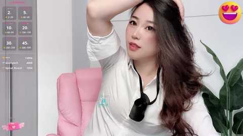 Media: Video of an East Asian woman with long, wavy black hair, wearing a white blouse, black necklace, and headphones, sitting in a pink office chair, with a calendar and potted plant in the background.