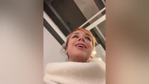 Media: Video of a smiling, fair-skinned woman with blonde hair, wearing a white sweater, viewed from below, indoors with a modern ceiling.