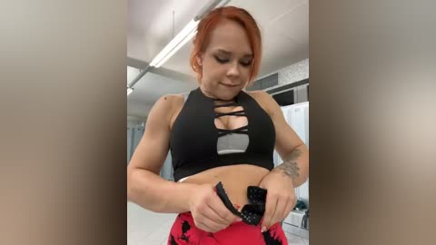 Media: Video of a fair-skinned, red-haired woman with a tattooed arm wearing a black crop top with cut-out details, adjusting her red and black patterned pants in a modern, brightly lit room with white walls and ceiling.