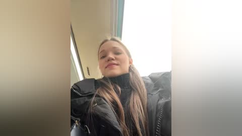 Media: Video of a young woman with long, straight brown hair, fair skin, and wearing a black puffy jacket, standing in a brightly lit room with a window behind her.