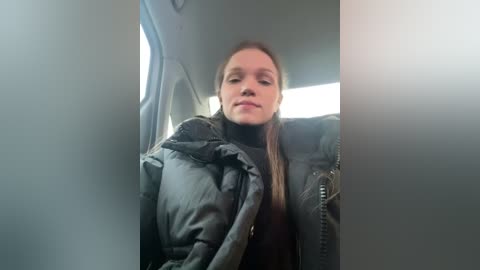 Media: Video of a young Caucasian woman with light skin and long, straight brown hair, wearing a black puffy jacket, sitting in a car with a grey interior.