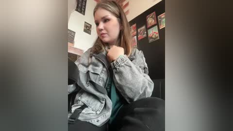 Media: Video of a young woman with light skin and straight brown hair, wearing a gray denim jacket over a green shirt, seated indoors. Background includes framed posters and a blackboard.