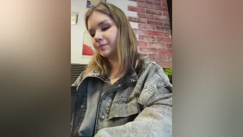 Media: Video of a young woman with light skin and shoulder-length blonde hair, wearing a denim jacket and a white shirt, standing against a brick wall and a red abstract painting in an indoor setting.