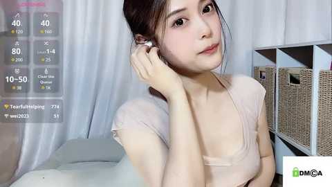 Media: A video of a young Asian woman with fair skin and dark hair, wearing a light pink top, in a cozy bedroom with white curtains and wicker baskets.