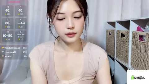 Media: A video of an Asian woman with fair skin, dark hair, and a soft V-neck top, wearing white earbuds. The background features white curtains and beige woven storage bins.